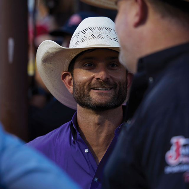 Dustin Boquet wearing a straw hat and a purple shirt talking to another man in a black shirt.